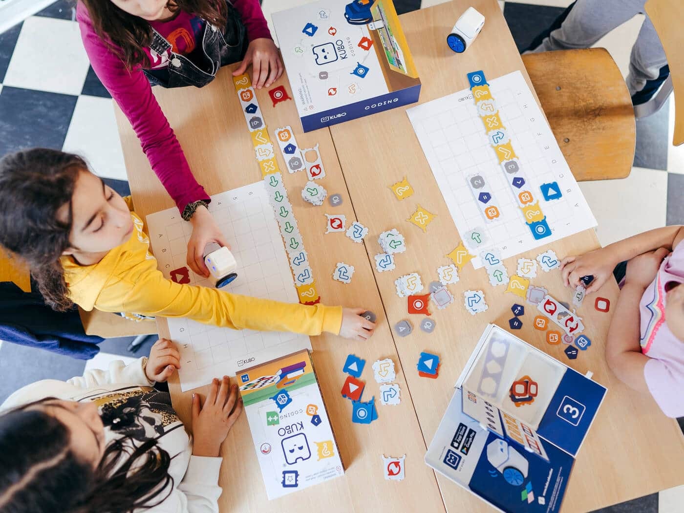 Three students at a classroom table snapping TagTiles into routes on activity maps, with the KUBO robot and the Starter Set box
